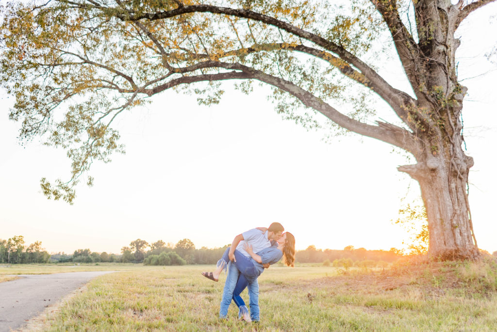 The groom is dipping the bride and giving her a kiss under a beautiful tree during a fall golden hour engagement session.