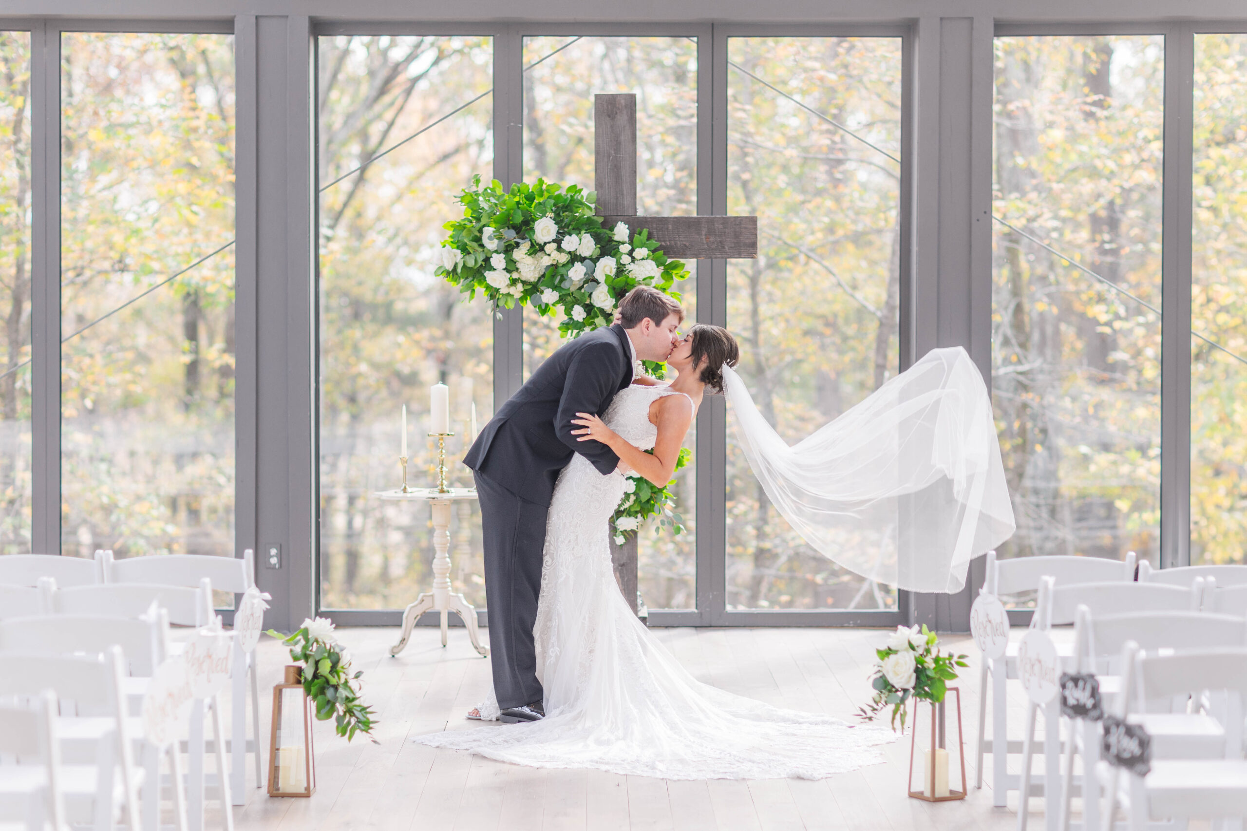 Groom dipping his bride back and kissing her in front of a wooden cross with greenery and white roses inside a luxurious glass chapel by Nashville wedding photographer, Brooke Elliott